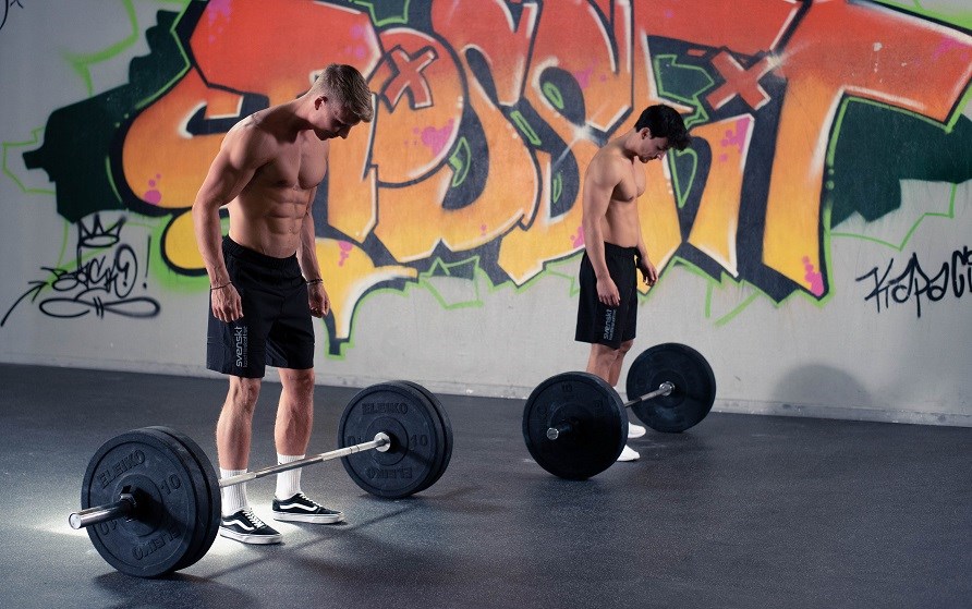 Två män inne på ett gym står bakom varsin skivstång lastad med vikter.