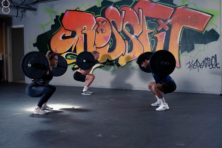 En kvinna och två  män på ett gym som tränar knäböj med varsin stång på ryggen,