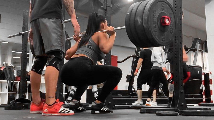 Bild på kvinna på ett gym som utför övningen knäböj med skivstång på ryggen.