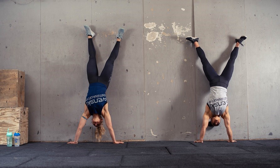Två kvinnor står på händer mot en vägg inne på ett gym.