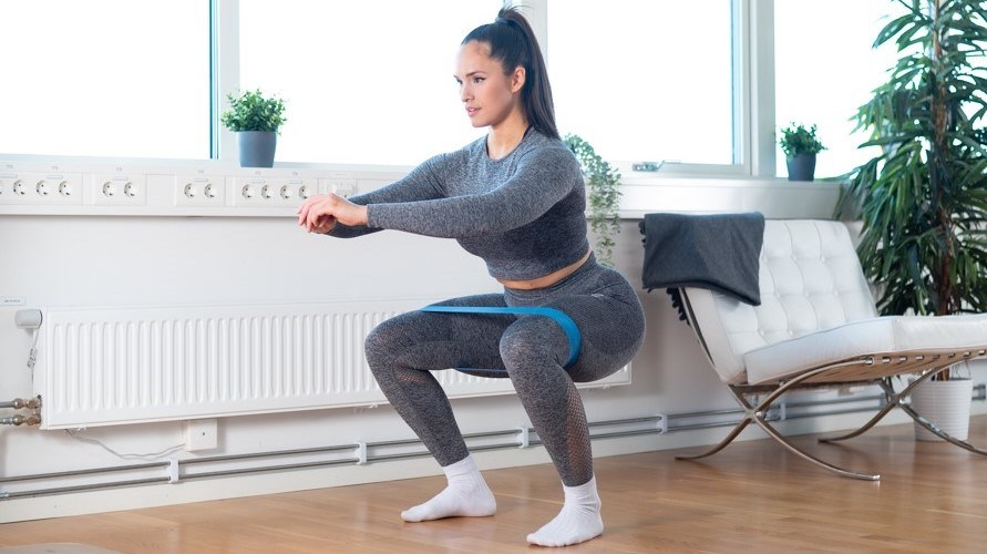 Kvinna gör övningen knäböj med gummiband runt benen.