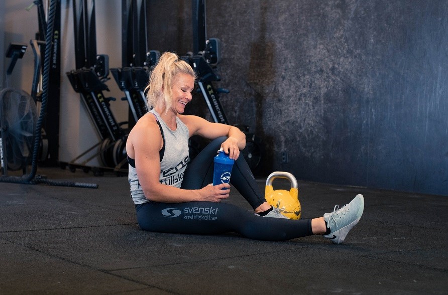 Kvinna på gym sitter på golvet med en shaker i handen.