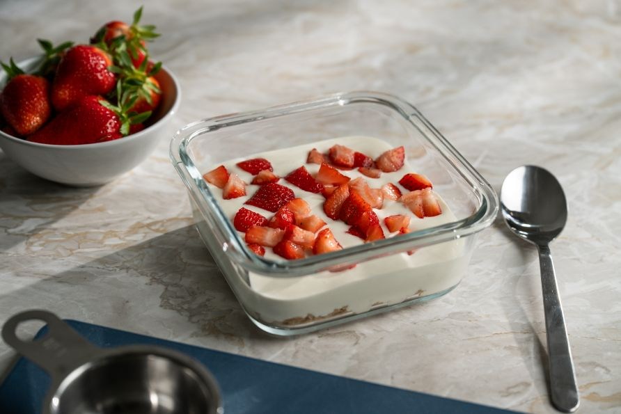 Glasmatl�da med proteinrik citron- och vaniljcheesecake som st�r p� ett bord tillsammans med en sked och en sk�l med jordgubbar