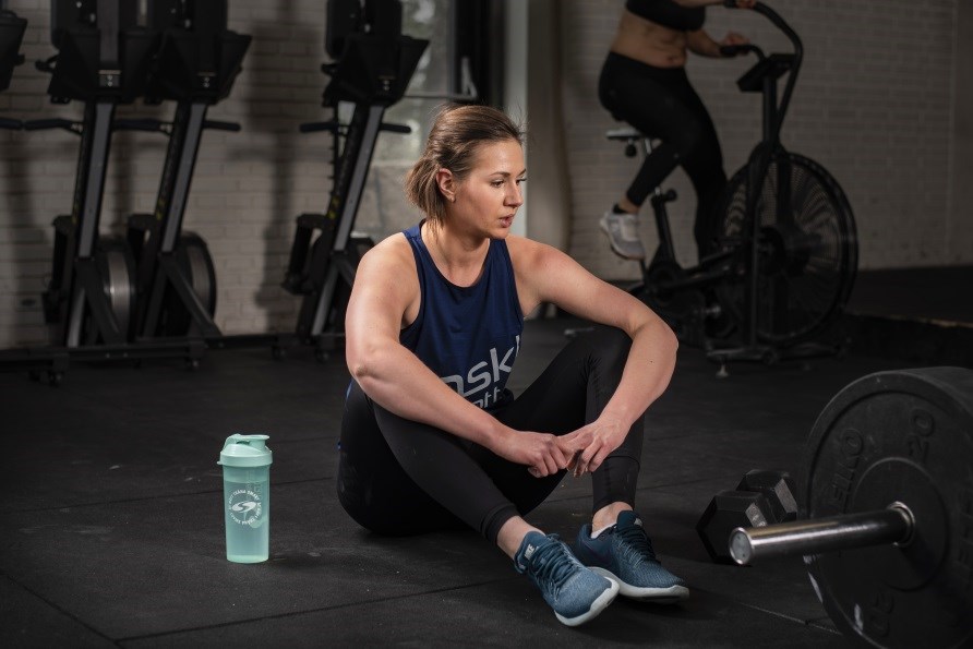 Kvinna sitter på golvet på ett gym, med en shaker bredvid sig. 