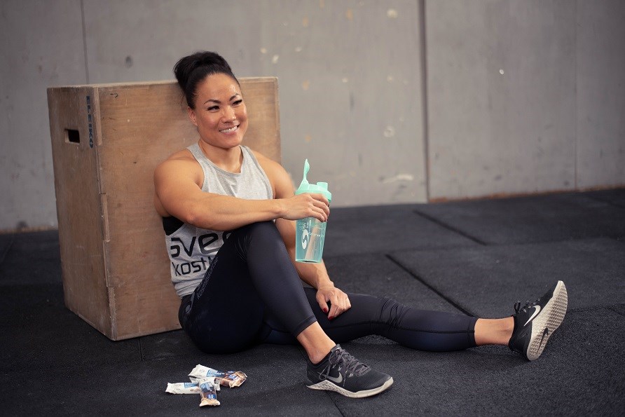 Kvinna som sitter lutad mot en box inne på ett gym. Håller en grön shaker från Svenskt Kosttillskott i handen.